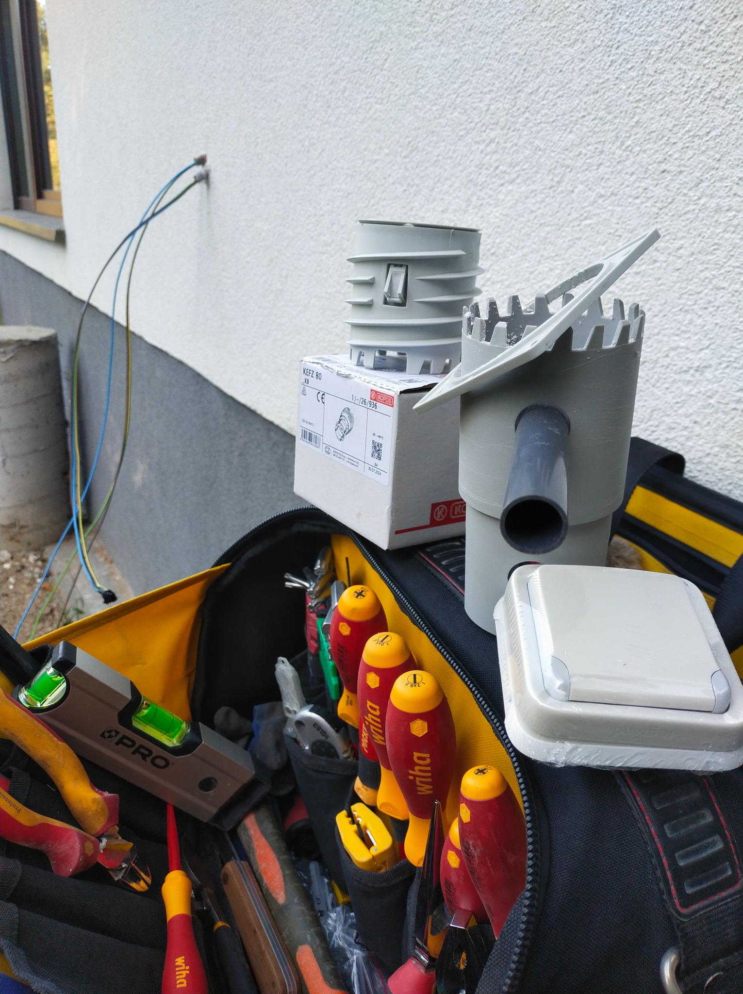 Otwarty torba narzędziowa z poziomnicą i różnymi śrubokrętami marki Wiha, obok puszka instalacyjna KEFZ 80 KB i elementy instalacji odgromowej na tle ściany budynku z wystającymi przewodami.