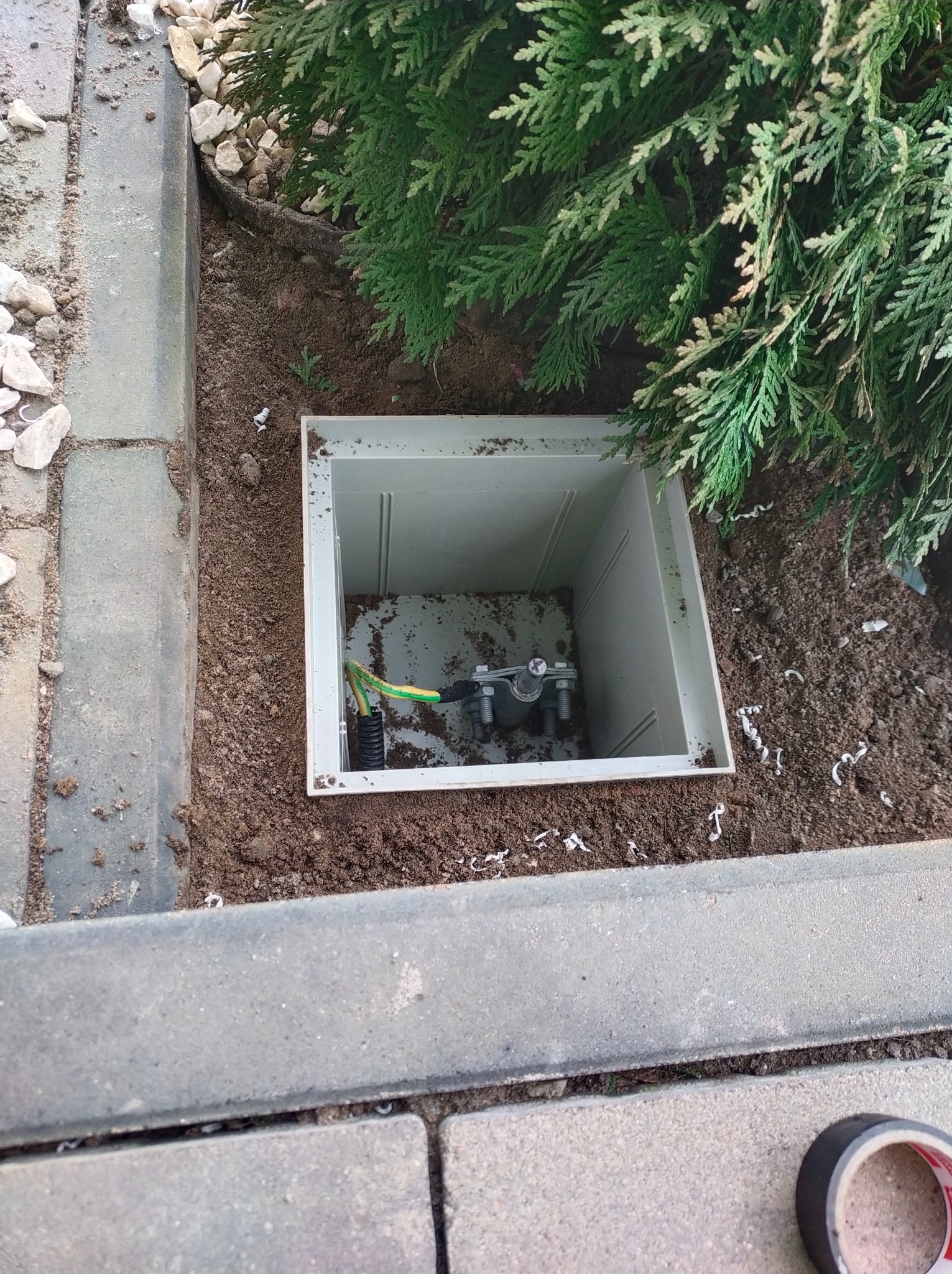 Złącze uziemiające w plastikowej studzience rewizyjnej, umieszczone w ziemi obok niskiego żywopłotu z tui, widok z góry.