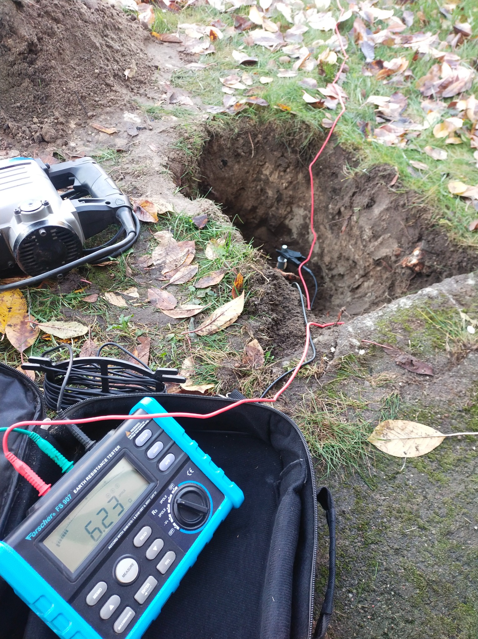 Miernik rezystancji uziemienia Forscher FS 907 z przewodami podłączonymi do wbitych w ziemię elektrod, obok wykopany dół i młot udarowy na trawie z liśćmi.