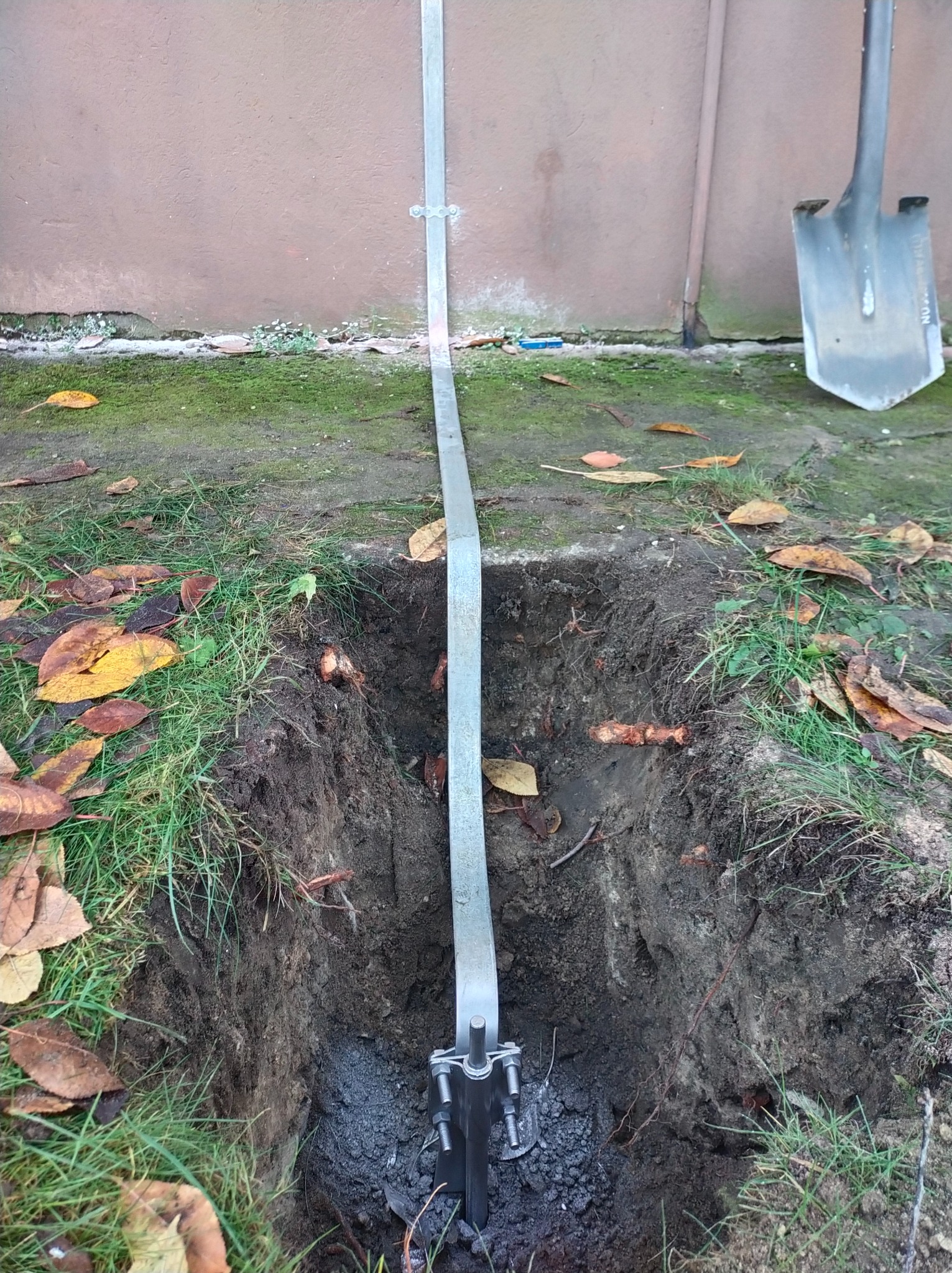 Uziemienie pionowe z bednarką w wykopie, widoczna łopata, jesienne liście na trawie.