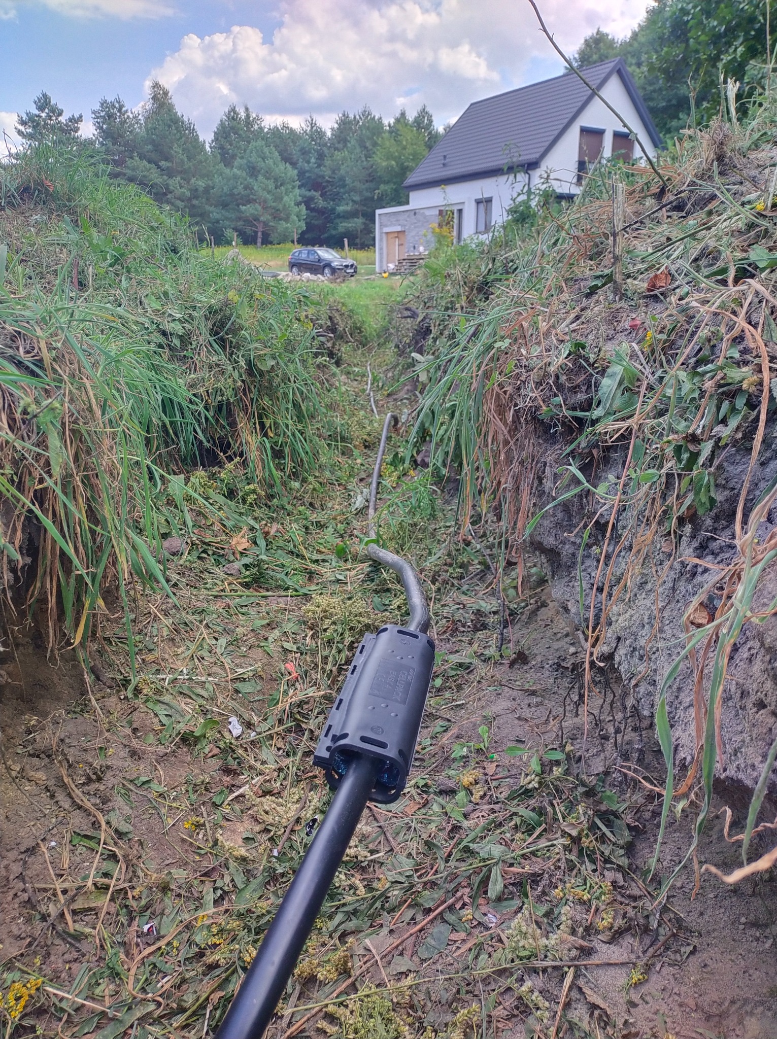 Kabel elektryczny zakopany w wykopie na tle domu jednorodzinnego i lasu, widoczne świeże ślady wykopu i luźna ziemia.