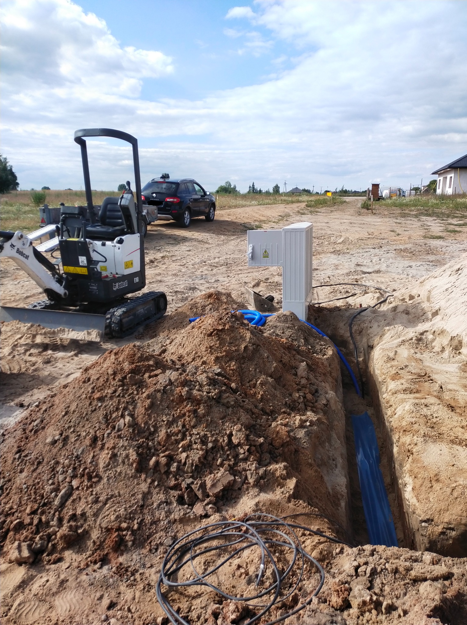 Wykonanie wykopu pod przyłącze elektryczne; widoczna skrzynka elektryczna, rury ochronne i mini koparka Bobcat E10 w piaszczystym terenie.