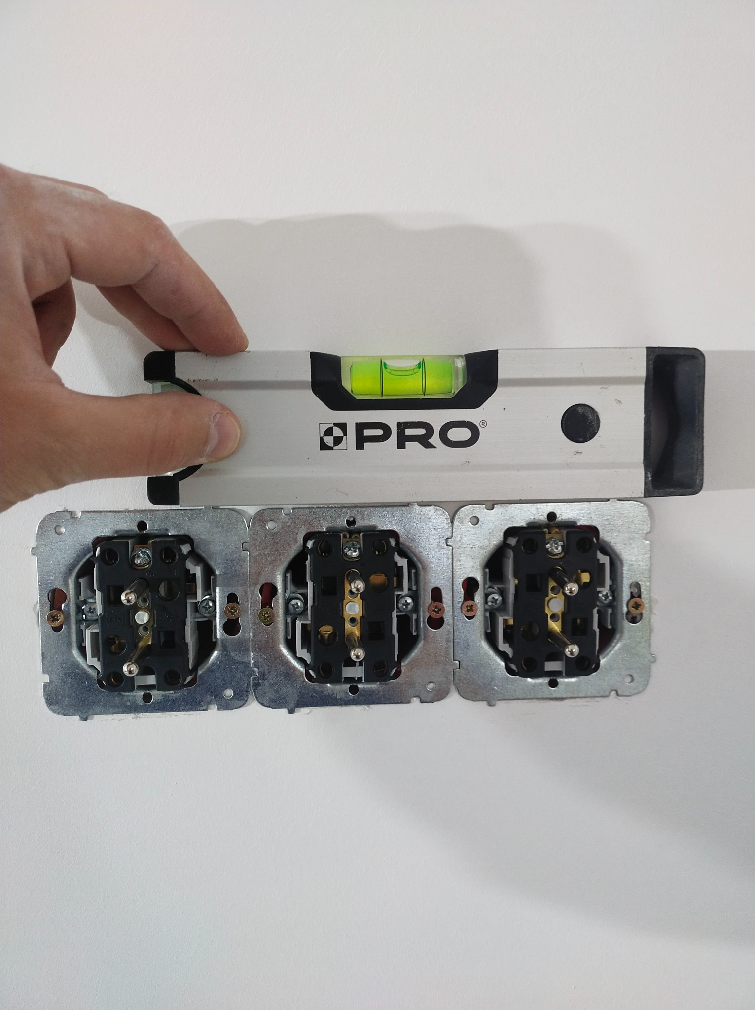 Poziomica PRO przykładana do potrójnego gniazdka elektrycznego w trakcie montażu, widoczne przewody i elementy mocujące.