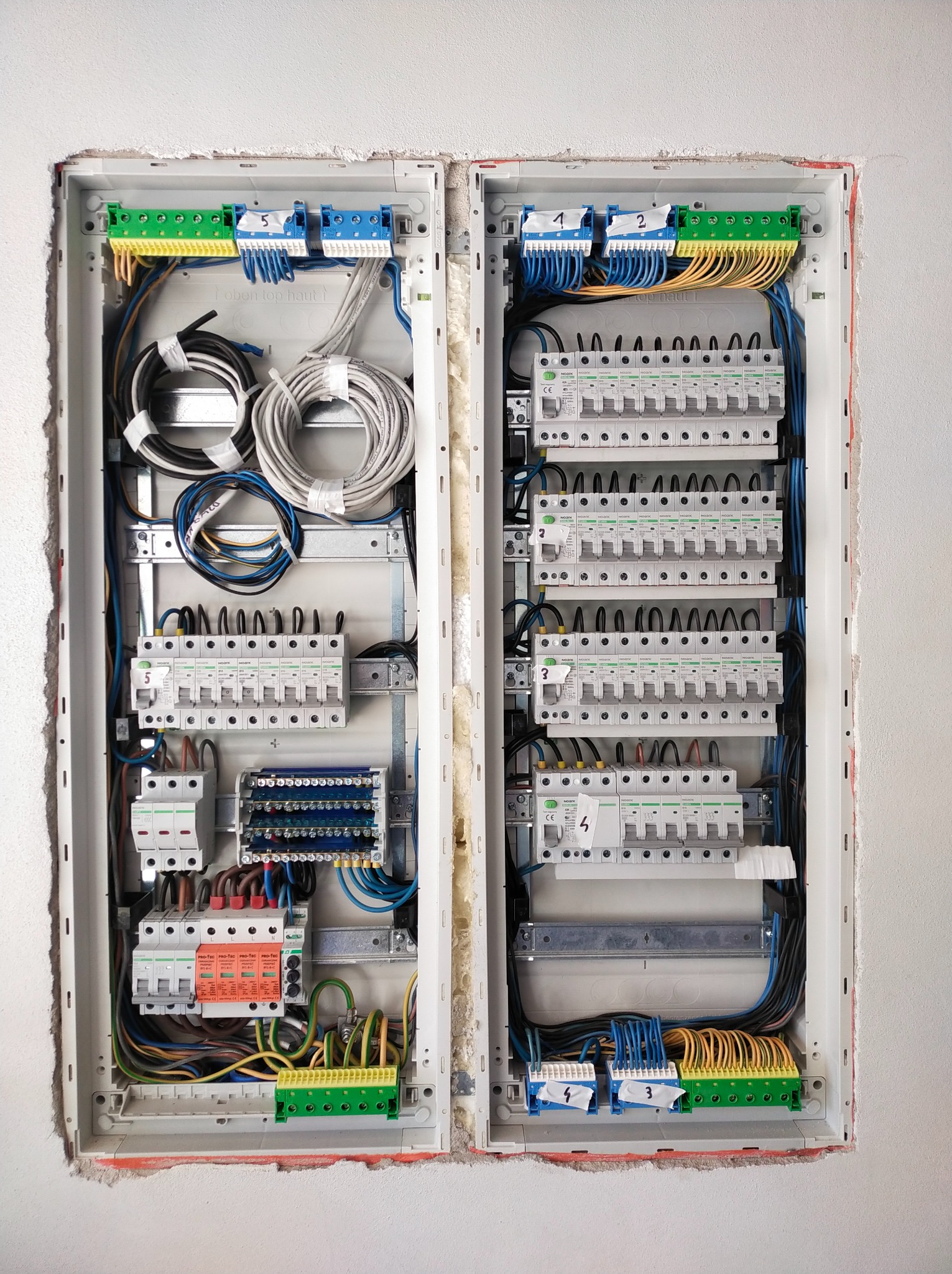 Dwie rozdzielnice elektryczne wnękowe, widoczne liczne przewody, bezpieczniki, listwy zaciskowe; instalacja w trakcie montażu, ściana wokół wnęk nie wykończona.