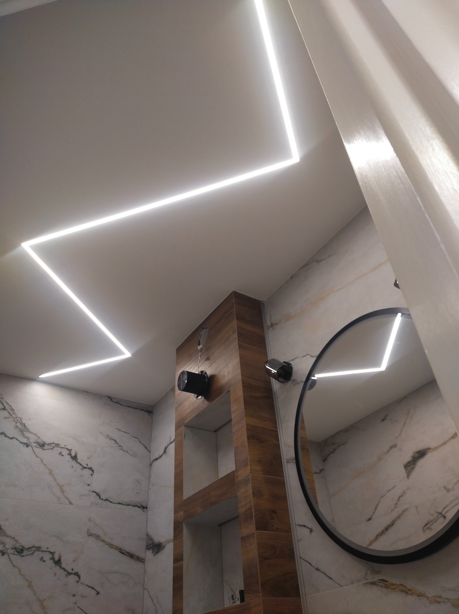 Nowoczesna łazienka z geometrycznym oświetleniem LED na suficie, marmurowe płytki na ścianach, okrągłe lustro i drewniane półki w zabudowie.