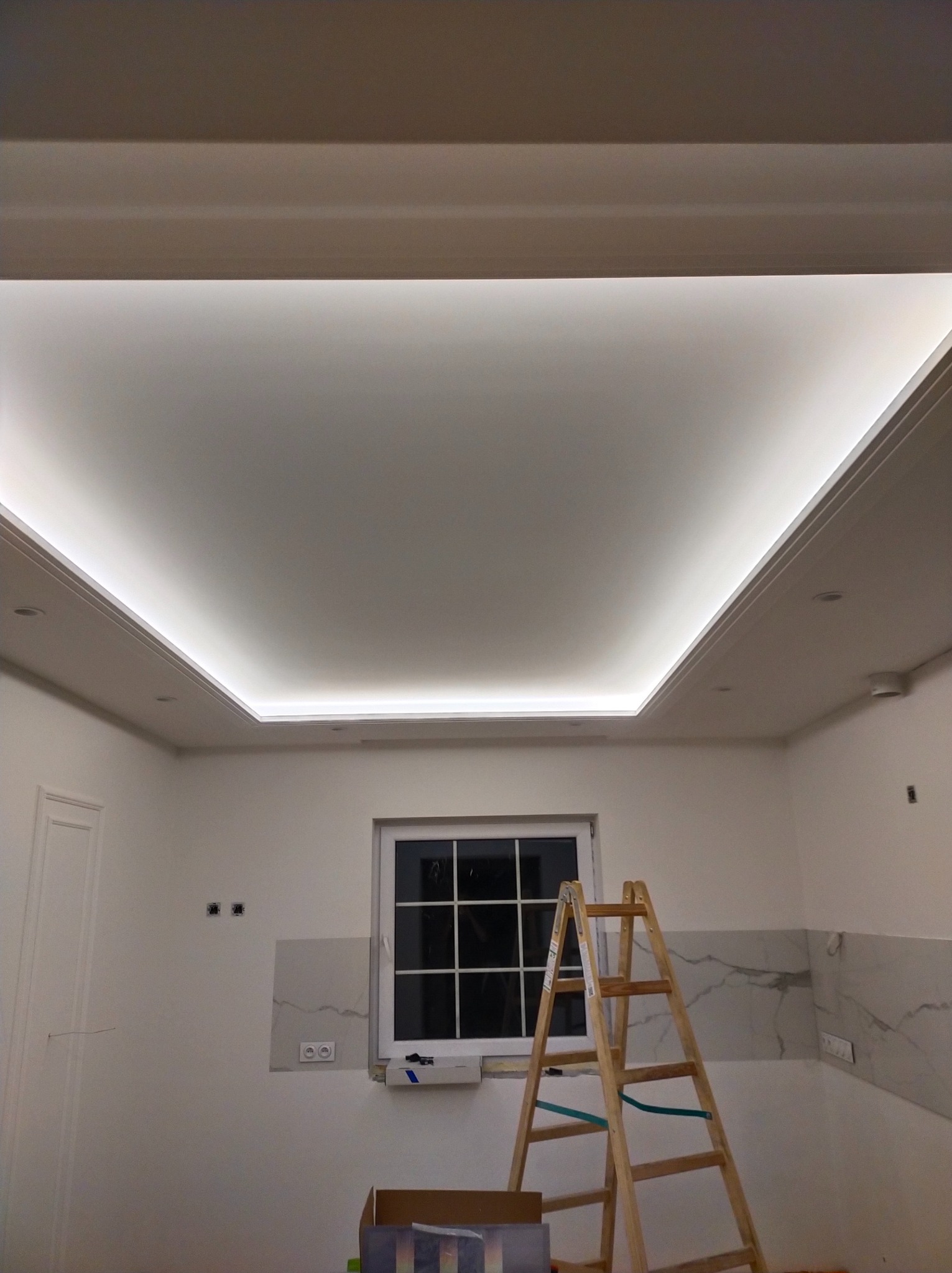 Podwieszany sufit z oświetleniem LED w trakcie montażu, widoczna drewniana drabina i przygotowane gniazdka elektryczne na ścianie.