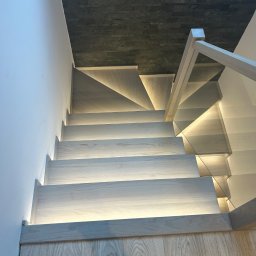 Milano - Nowoczesne schody drewniane z podświetleniem LED, szklana balustrada, widok z góry na stopnie z jasnego drewna i ciemną ścianę z kamienia dekoracyjnego.