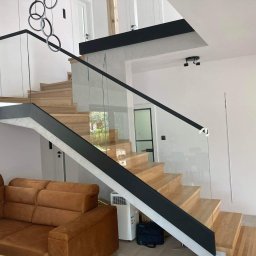 Milano - Nowoczesne schody drewniane z balustradą ze szkła, czarne elementy wykończeniowe, widoczny fragment brązowej sofy i designerskiej lampy sufitowej.