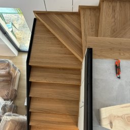 Milano - Kątowe ujęcie schodów z jasnego drewna z czarną balustradą, widoczne narzędzie do cięcia na szarej podłodze oraz zapakowane meble przy oknie.