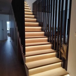Milano - Nowoczesne schody drewniane z podświetleniem LED na każdym stopniu, czarne pionowe barierki, widok z dołu na górę, jasne drewno, minimalistyczny design, przeszklona balustrada na górze.