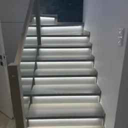 Nowoczesne schody drewniane z podświetleniem LED stopni, balustrada szklana, jasne drewno, ściana z dekoracyjnej cegły w tle.