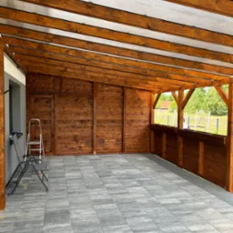 Zadaszony taras z drewnianą konstrukcją, wyłożony szarą kostką brukową, widoczna drabina oparta o ścianę budynku.