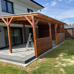 Milano - Zadaszony taras z drewnianą konstrukcją i częściowym obudowaniem z desek, z aluminiową drabiną opartą o filar, na tle nowoczesnego domu i trawnika