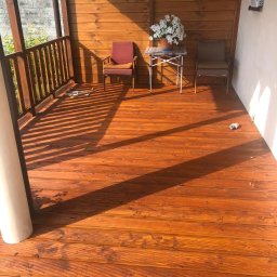 Milano - Zadaszony taras z drewnianą podłogą w odcieniach brązu, oświetlony promieniami słońca, z widocznymi dwoma krzesłami, stolikiem i kwiatami w doniczce.