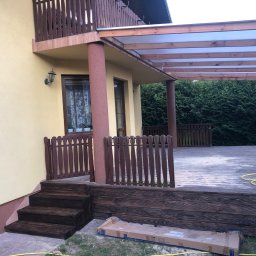 Milano - Drewniany taras z zadaszeniem przy domu w Żabiej Woli, z widocznymi schodami i fragmentem ogrodu. Częściowo pomalowany taras z widocznym wężem ogrodowym i kartonem po produkcie.