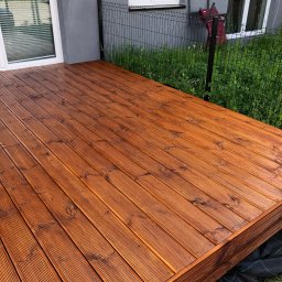 Milano - Nowy taras z desek ryflowanych, zabezpieczony lakierobejcą w odcieniu orzecha, widok z niskiej perspektywy, na tle szarej elewacji i zieleni.