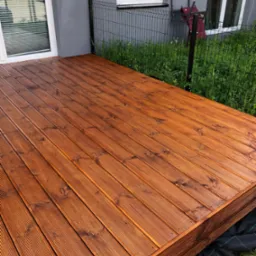 Nowy taras z desek ryflowanych, zabezpieczony lakierobejcą w odcieniu orzecha, widok z niskiej perspektywy, na tle szarej elewacji i zieleni.