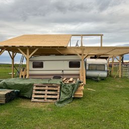 Milano - Budowa drewnianej wiaty z dachem z płyt OSB nad przyczepami kempingowymi, widoczne rusztowania, palety z drewnem i plandeka na trawie.