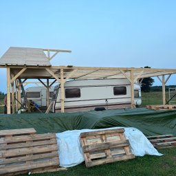 Milano - Budowa drewnianej konstrukcji z zadaszeniem obok przyczepy kempingowej, widoczne palety i plandeka ochronna w otoczeniu zielonego terenu.