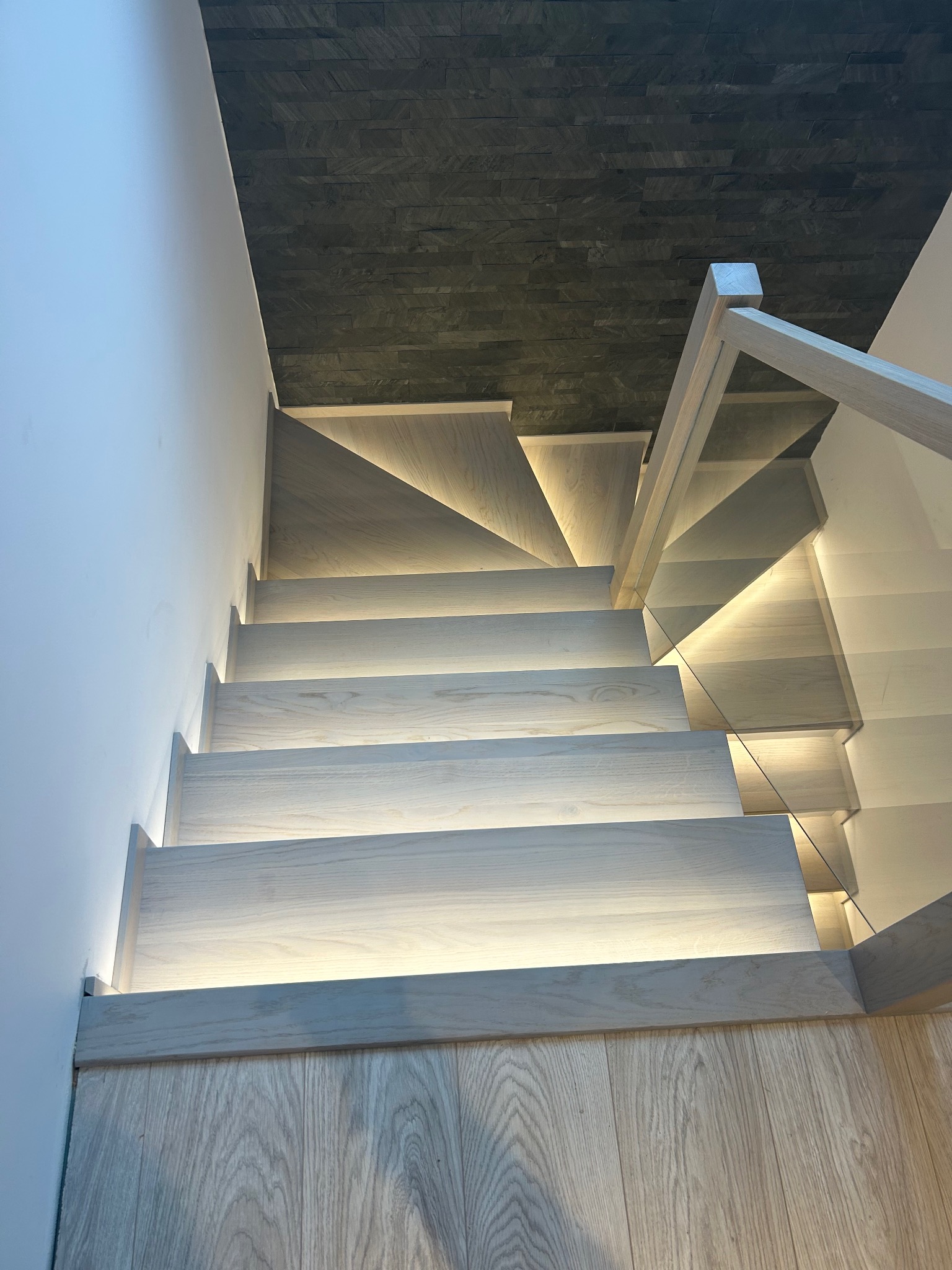 Nowoczesne schody drewniane z podświetleniem LED, szklana balustrada, widok z góry na stopnie z jasnego drewna i ciemną ścianę z kamienia dekoracyjnego.