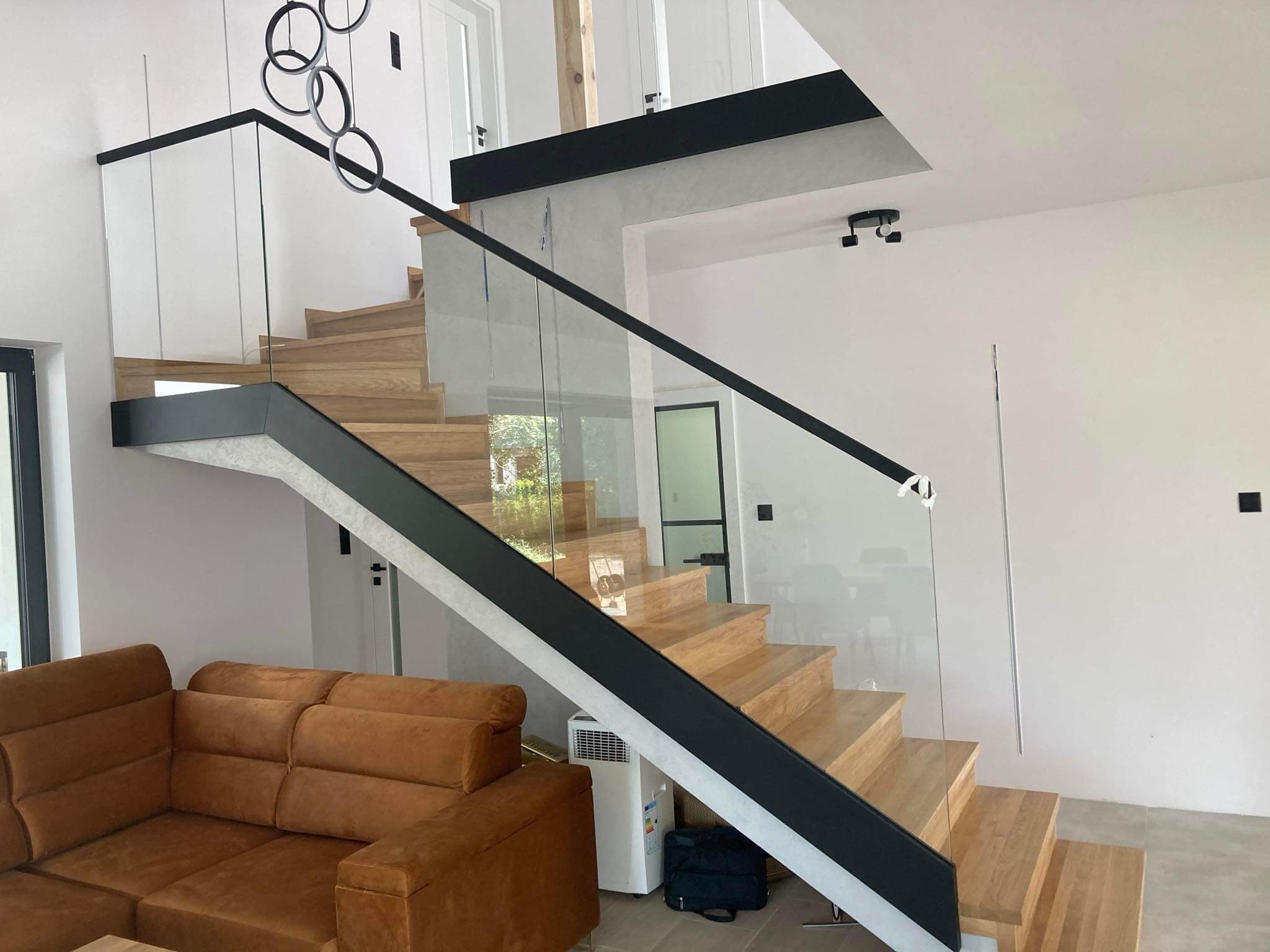 Nowoczesne schody drewniane z balustradą ze szkła, czarne elementy wykończeniowe, widoczny fragment brązowej sofy i designerskiej lampy sufitowej.