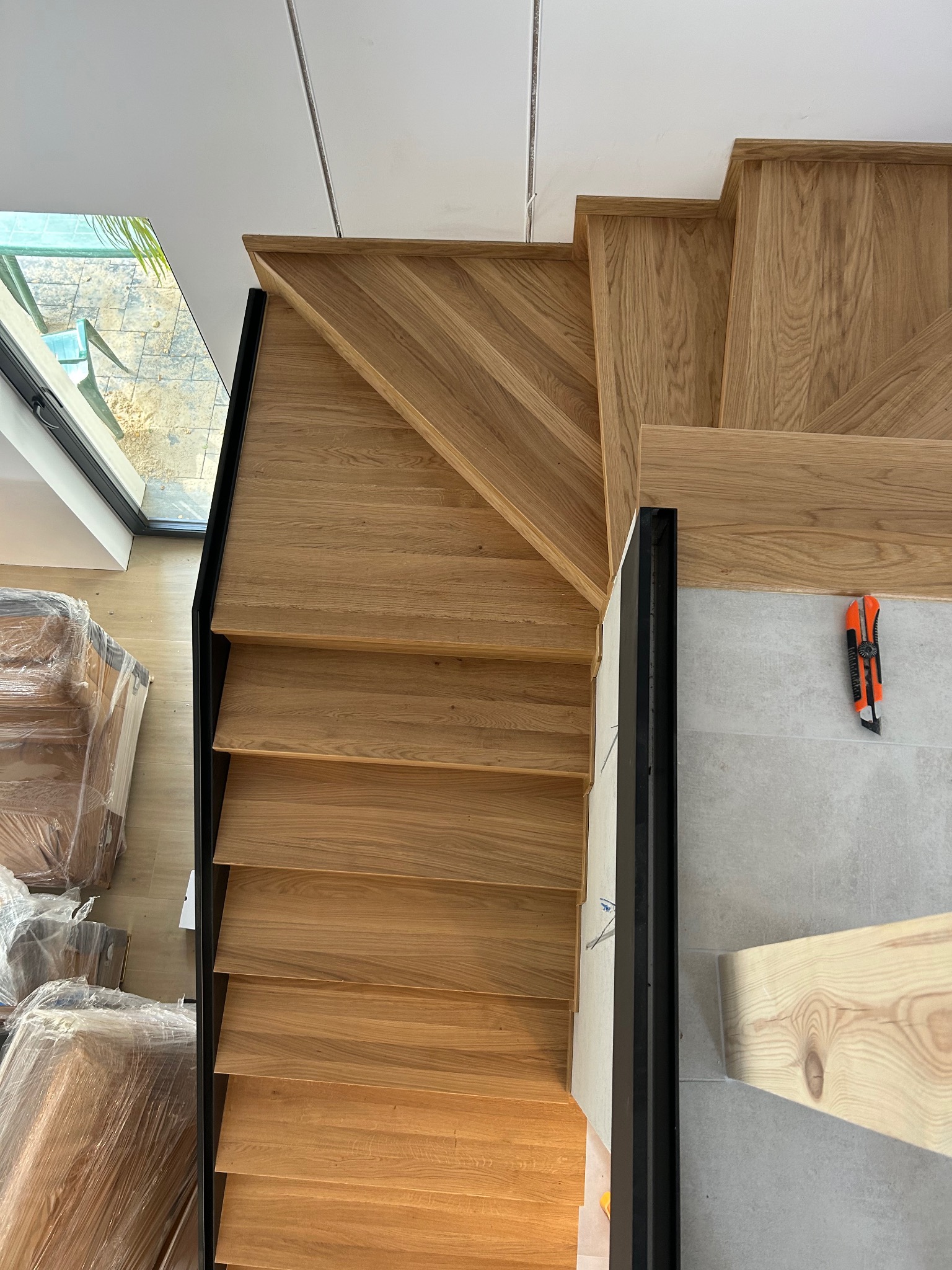 Kątowe ujęcie schodów z jasnego drewna z czarną balustradą, widoczne narzędzie do cięcia na szarej podłodze oraz zapakowane meble przy oknie.