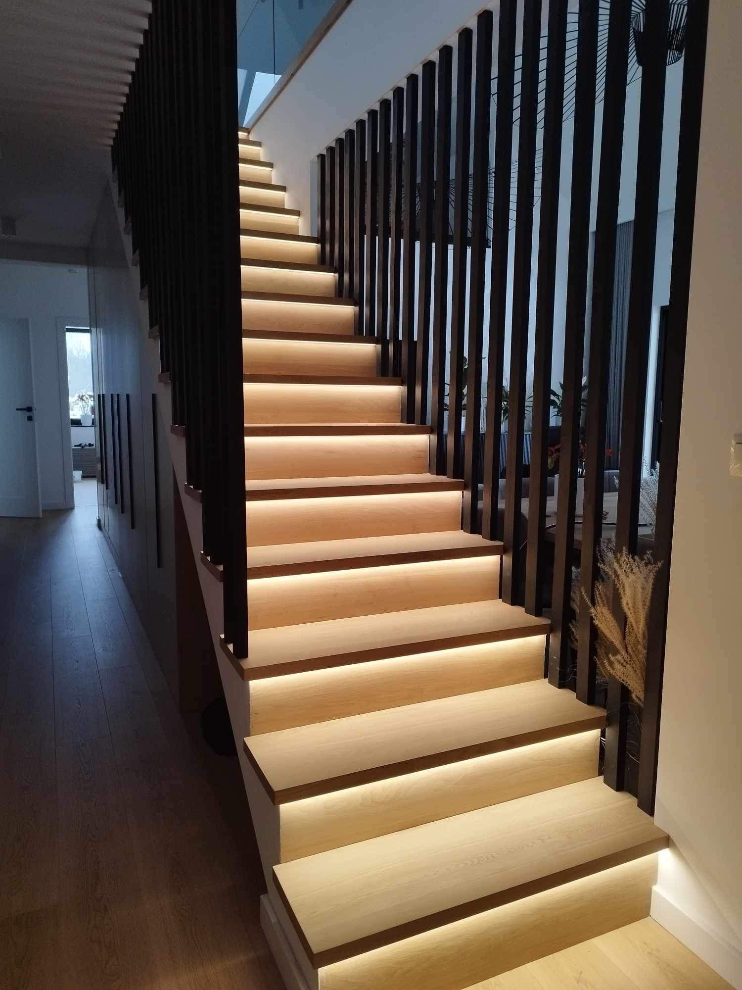 Nowoczesne schody drewniane z podświetleniem LED na każdym stopniu, czarne pionowe barierki, widok z dołu na górę, jasne drewno, minimalistyczny design, przeszklona balustrada na górze.
