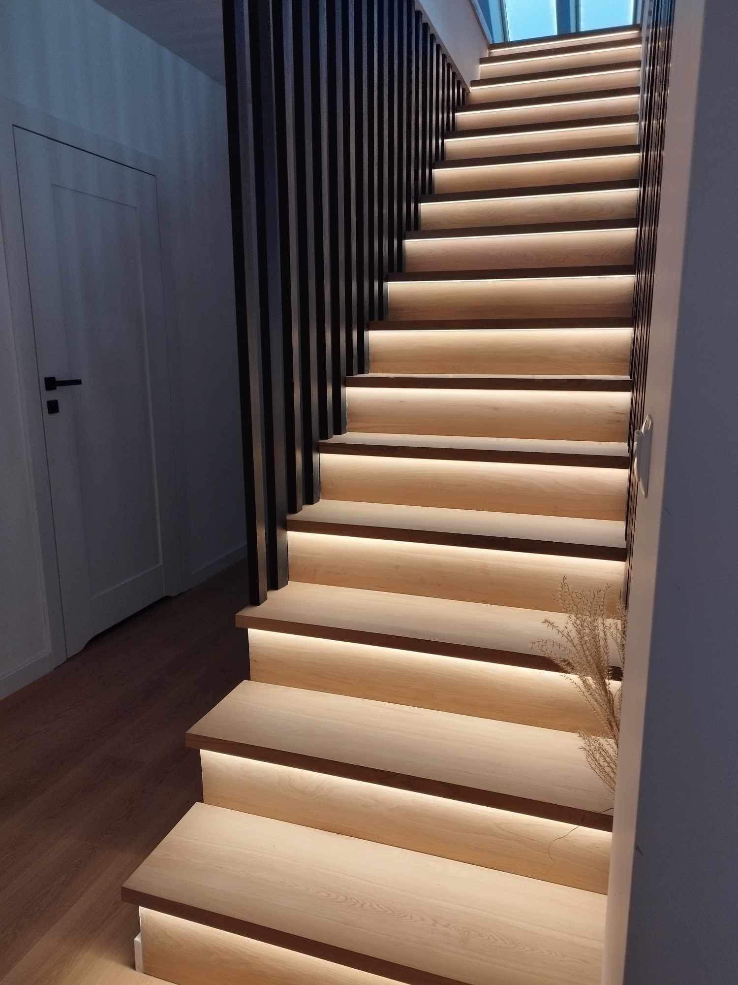 Nowoczesne, podświetlane schody drewniane z czarną balustradą, widok z dołu, oświetlenie LED podkreśla strukturę drewna i dodaje ciepła wnętrzu.