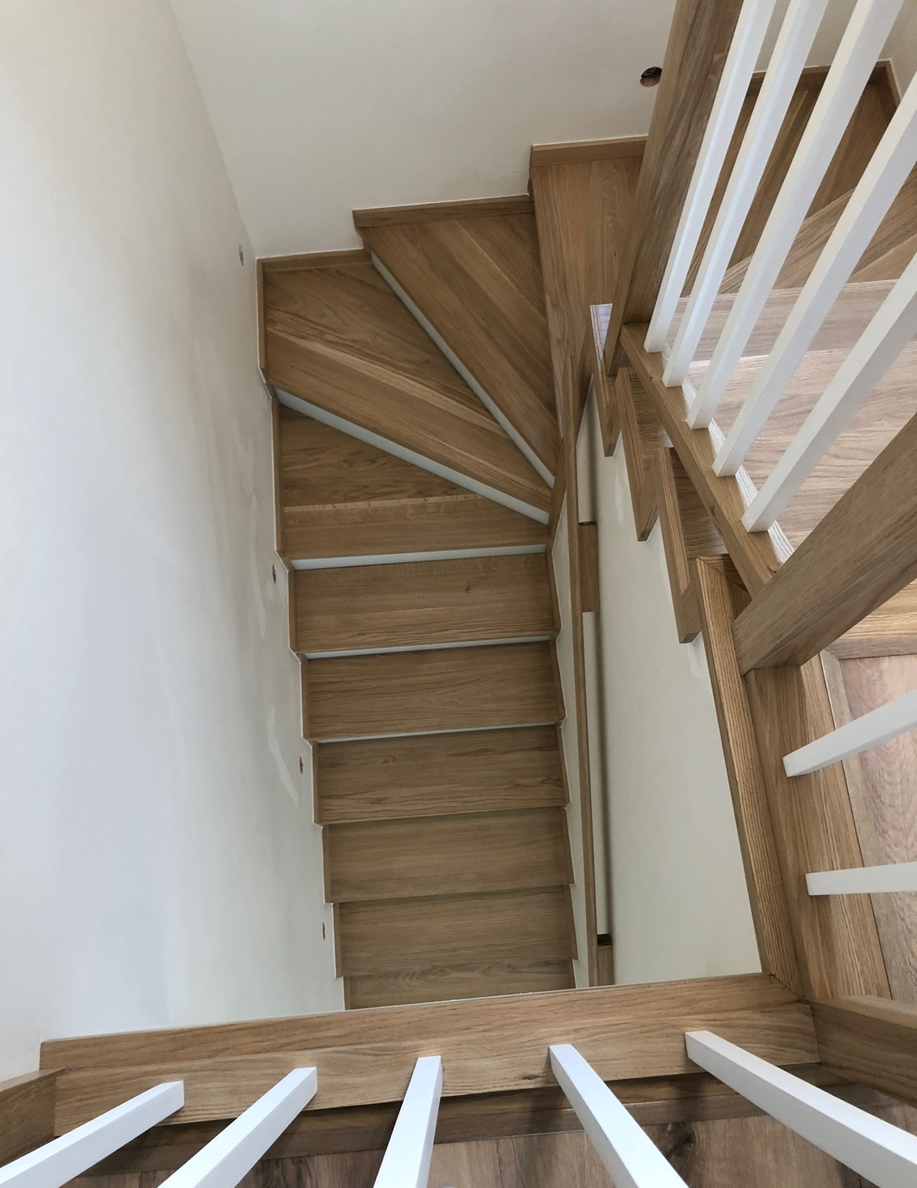 Widok z góry na drewniane schody z białymi balustradami, z widocznymi detalami usłojenia drewna i minimalistycznym wykończeniem ścian.
