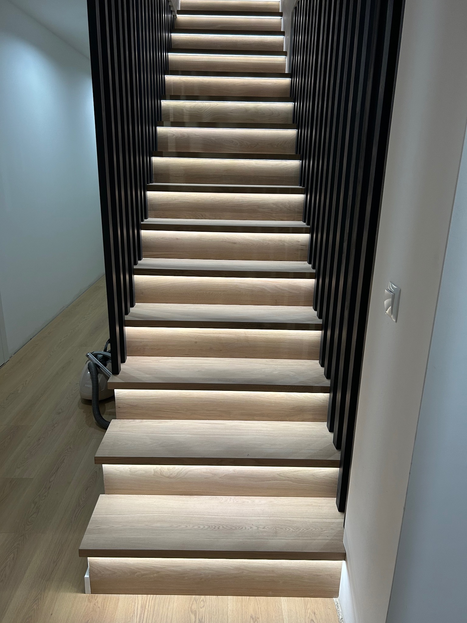 Nowoczesne schody drewniane z podświetleniem LED, czarne pionowe barierki, widoczny odkurzacz na podłodze.