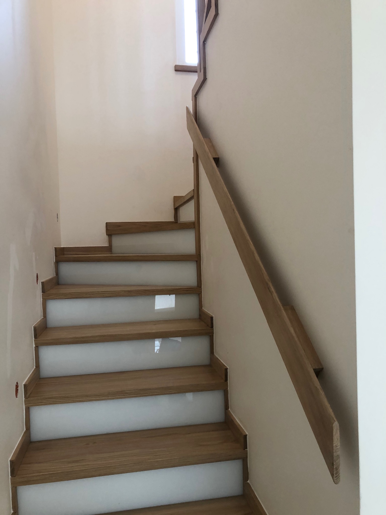 Nowoczesne schody z drewnianymi stopniami i białymi podstopnicami, drewniana balustrada przy ścianie, minimalistyczny design.