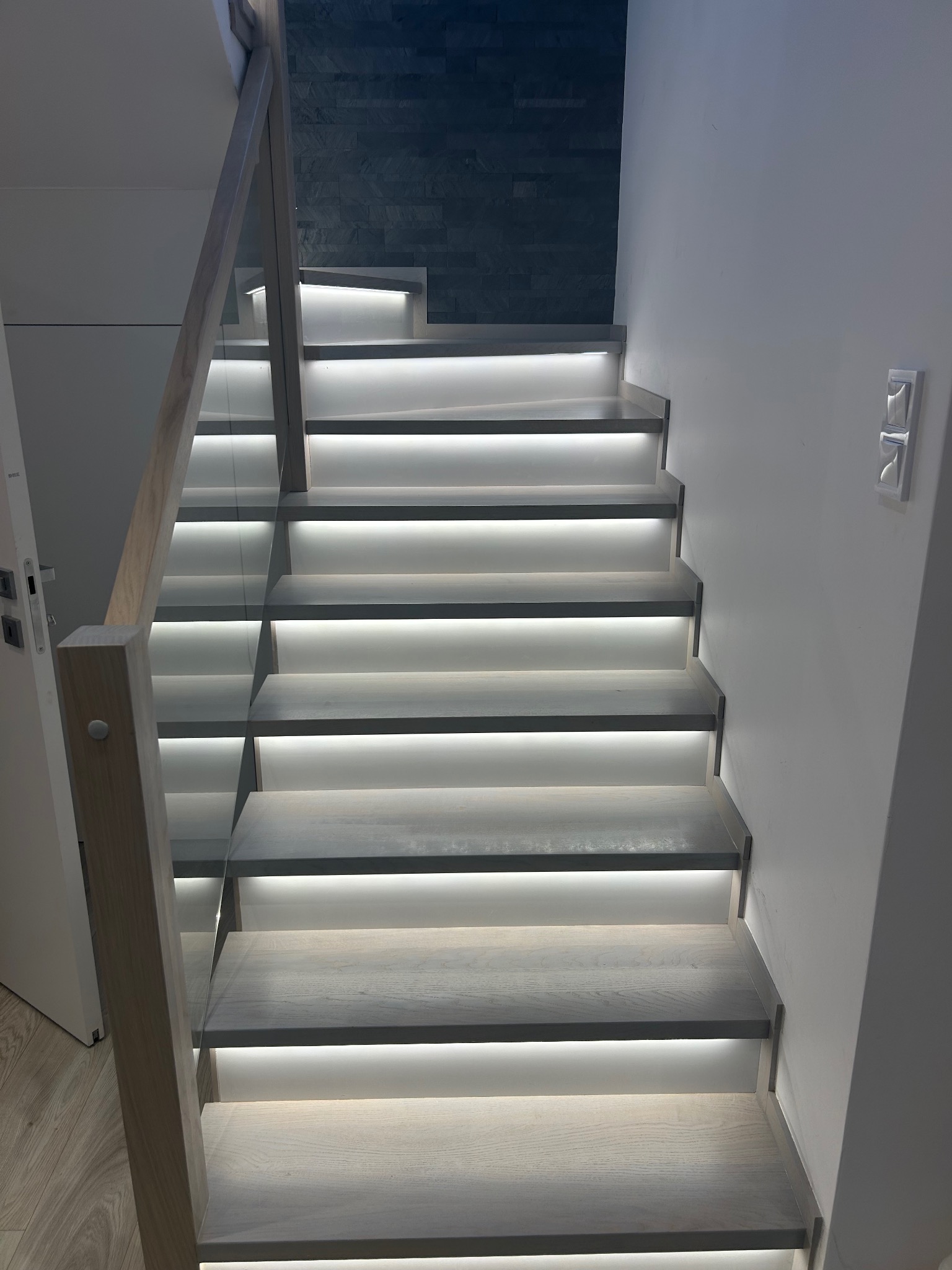 Nowoczesne schody drewniane z podświetleniem LED stopni, balustrada szklana, jasne drewno, ściana z dekoracyjnej cegły w tle.