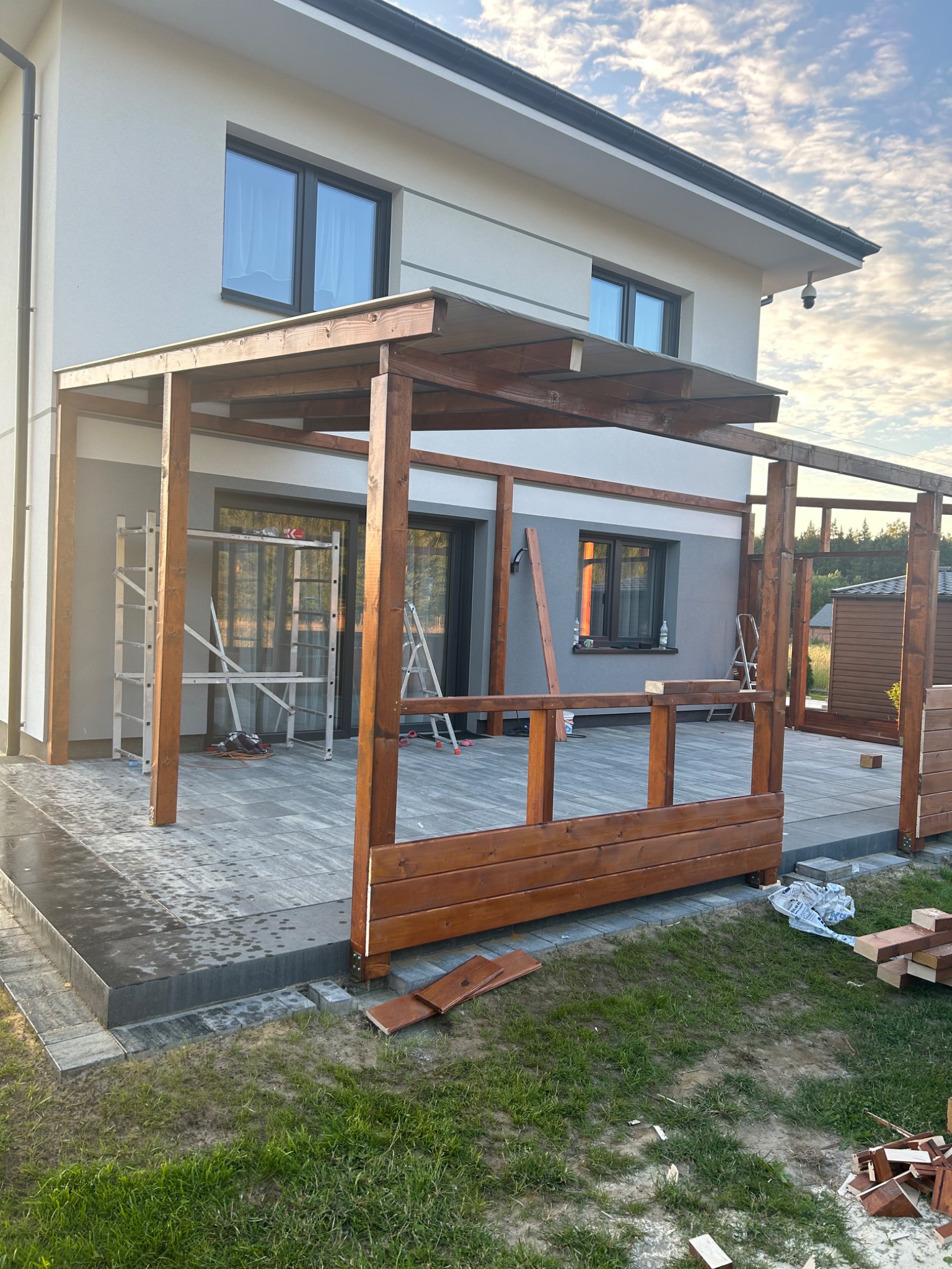 Budowa drewnianego tarasu przy domu jednorodzinnym, widoczne elementy konstrukcyjne, drabiny, narzędzia oraz pozostałości materiałów budowlanych na trawniku.