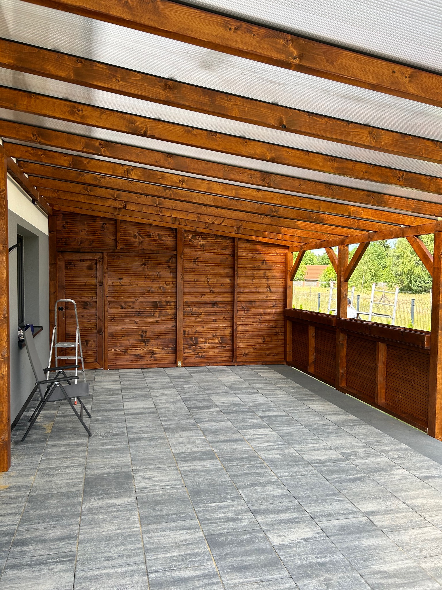 Zadaszony taras z drewnianą konstrukcją, wyłożony szarą kostką brukową, widoczna drabina oparta o ścianę budynku.