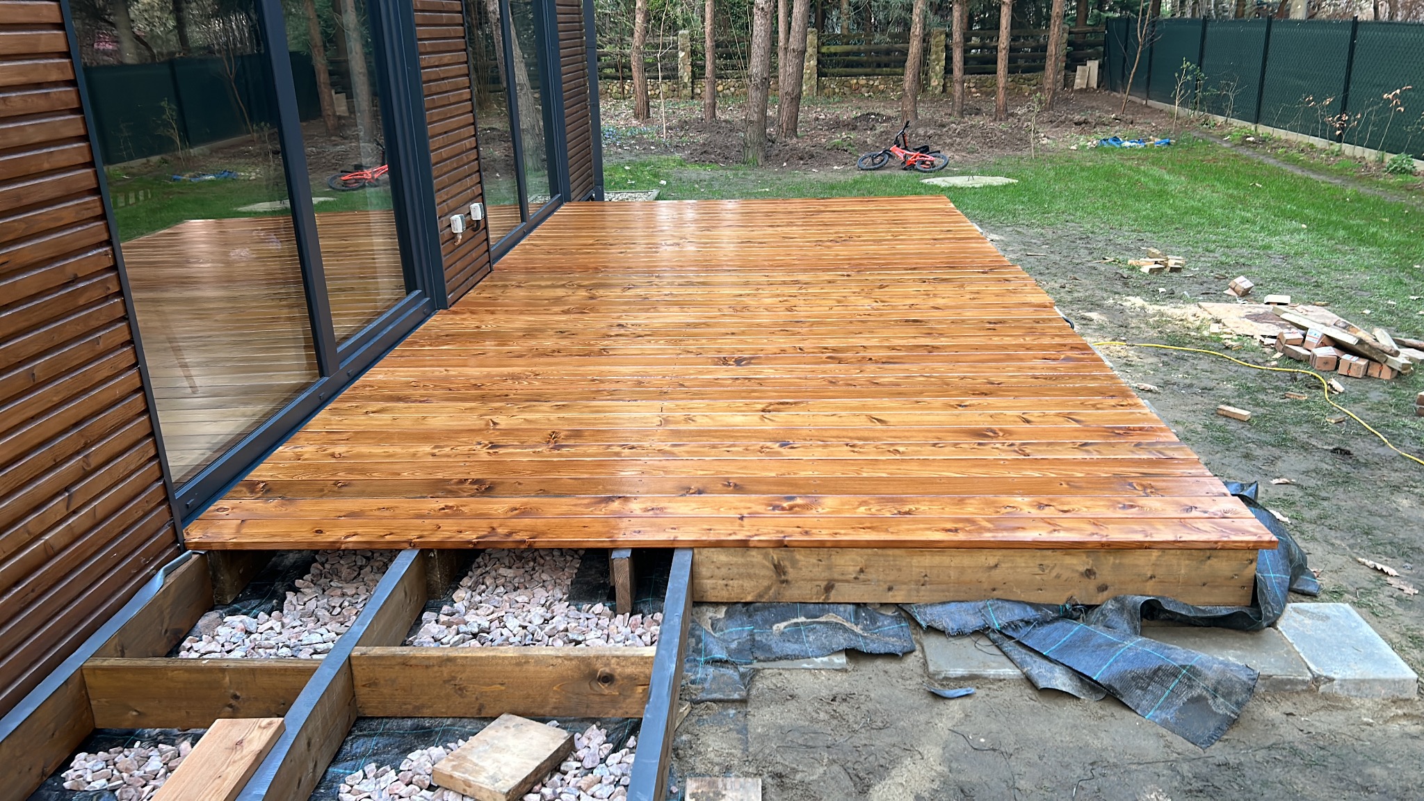 Nowo wybudowany taras drewniany z widoczną konstrukcją nośną wypełnioną kamieniami, świeżo polakierowane deski, tło: trawnik, drzewa i budynek z dużymi oknami.