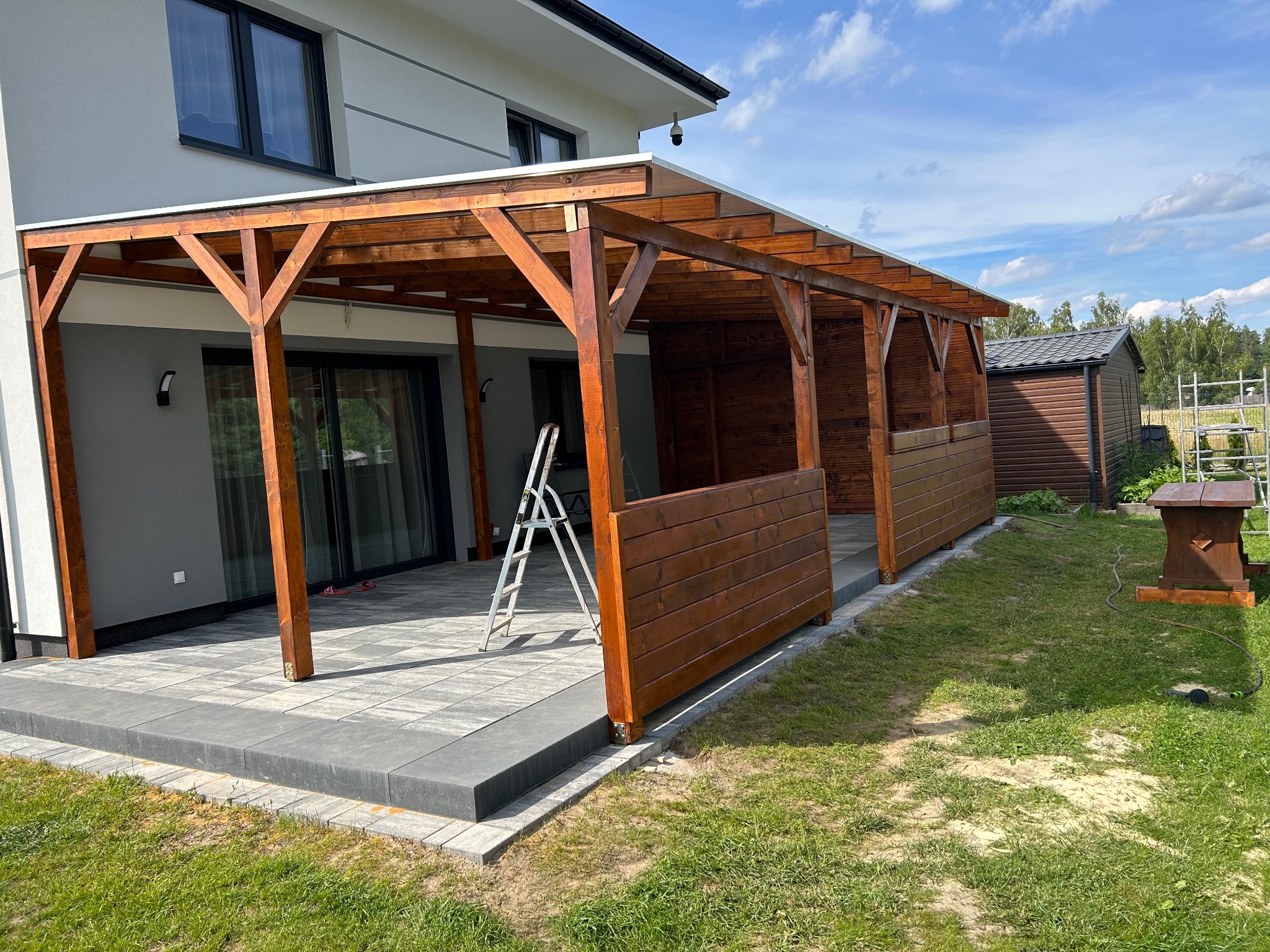 Zadaszony taras z drewnianą konstrukcją i częściowym obudowaniem z desek, z aluminiową drabiną opartą o filar, na tle nowoczesnego domu i trawnika