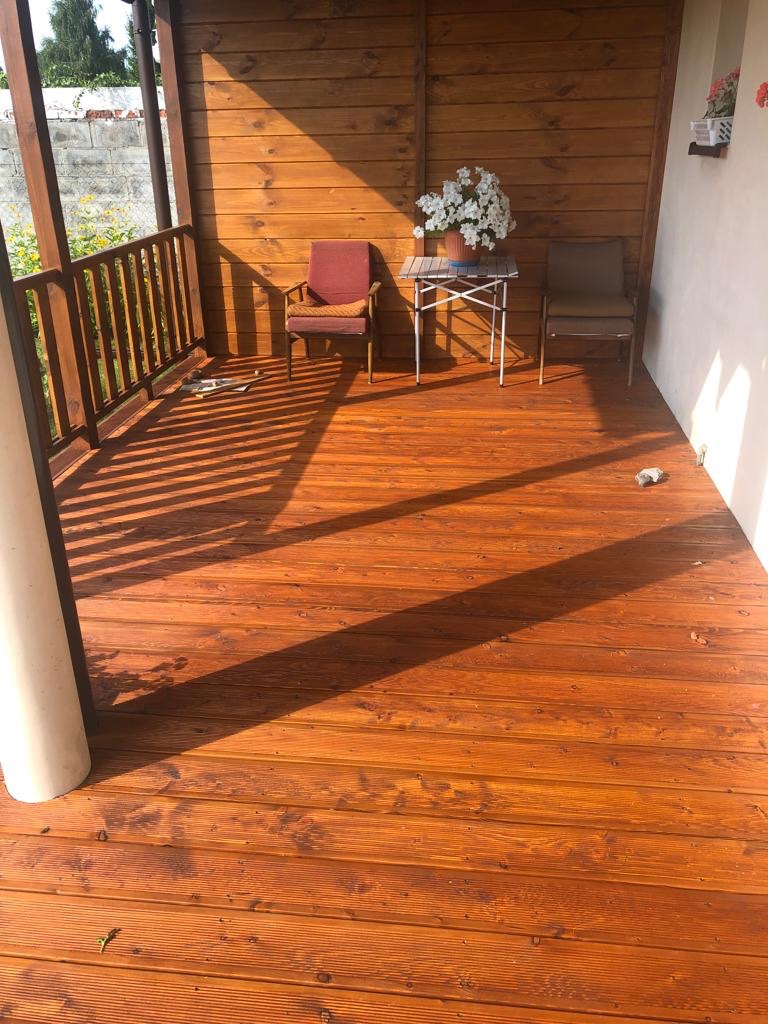 Zadaszony taras z drewnianą podłogą w odcieniach brązu, oświetlony promieniami słońca, z widocznymi dwoma krzesłami, stolikiem i kwiatami w doniczce.