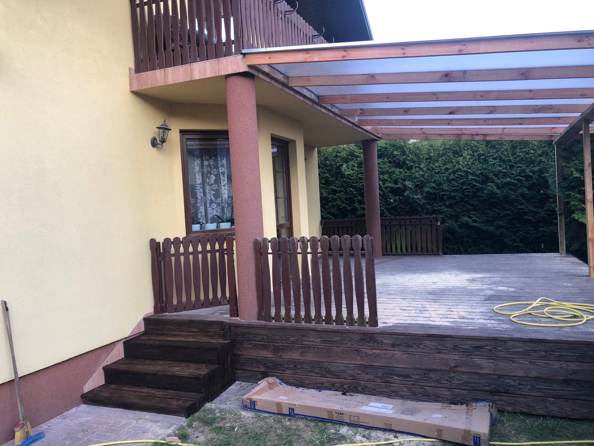 Drewniany taras z zadaszeniem przy domu w Żabiej Woli, z widocznymi schodami i fragmentem ogrodu. Częściowo pomalowany taras z widocznym wężem ogrodowym i kartonem po produkcie.