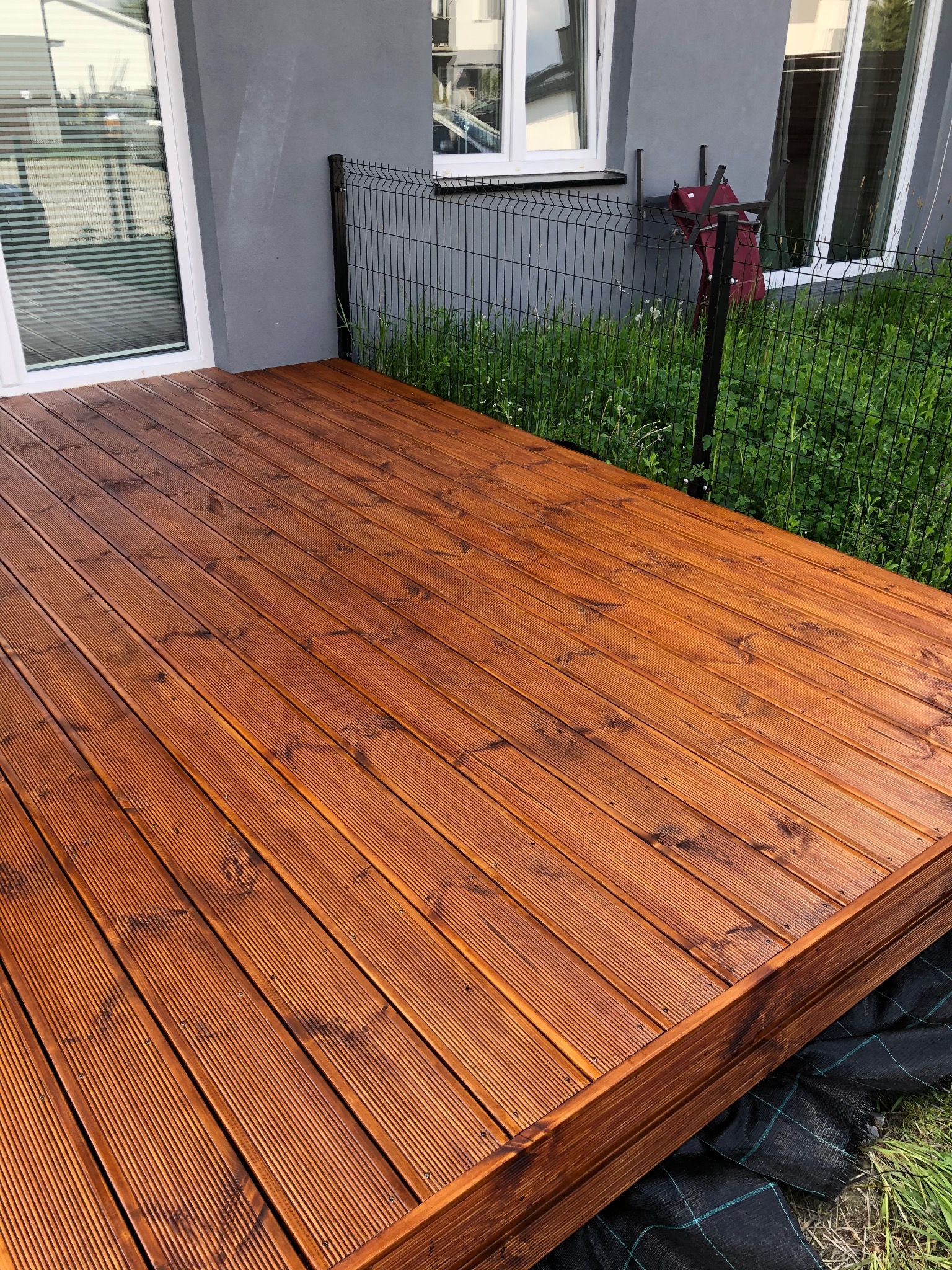 Nowy taras z desek ryflowanych, zabezpieczony lakierobejcą w odcieniu orzecha, widok z niskiej perspektywy, na tle szarej elewacji i zieleni.