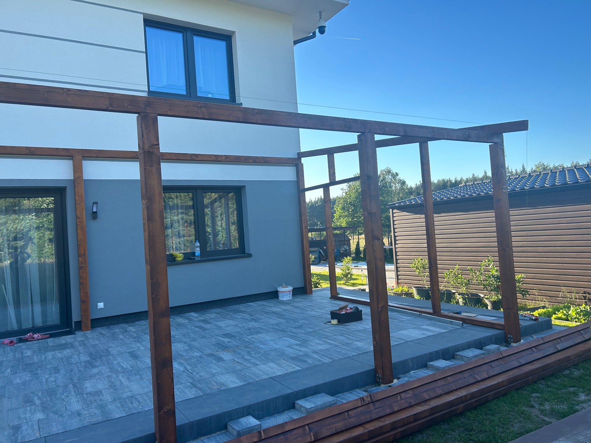 Konstrukcja drewnianej pergoli w trakcie montażu na tarasie wyłożonym szarą kostką brukową, widoczne narzędzia i elementy konstrukcyjne, tło stanowi nowoczesny dom i ogrodzenie.