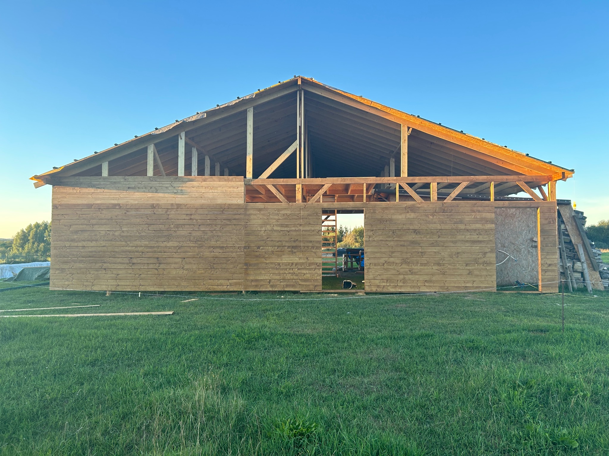 Wznoszona drewniana konstrukcja budynku gospodarczego z widocznym szkieletem dachu i częściowo obłożonymi ścianami na tle zielonego trawnika i błękitnego nieba.