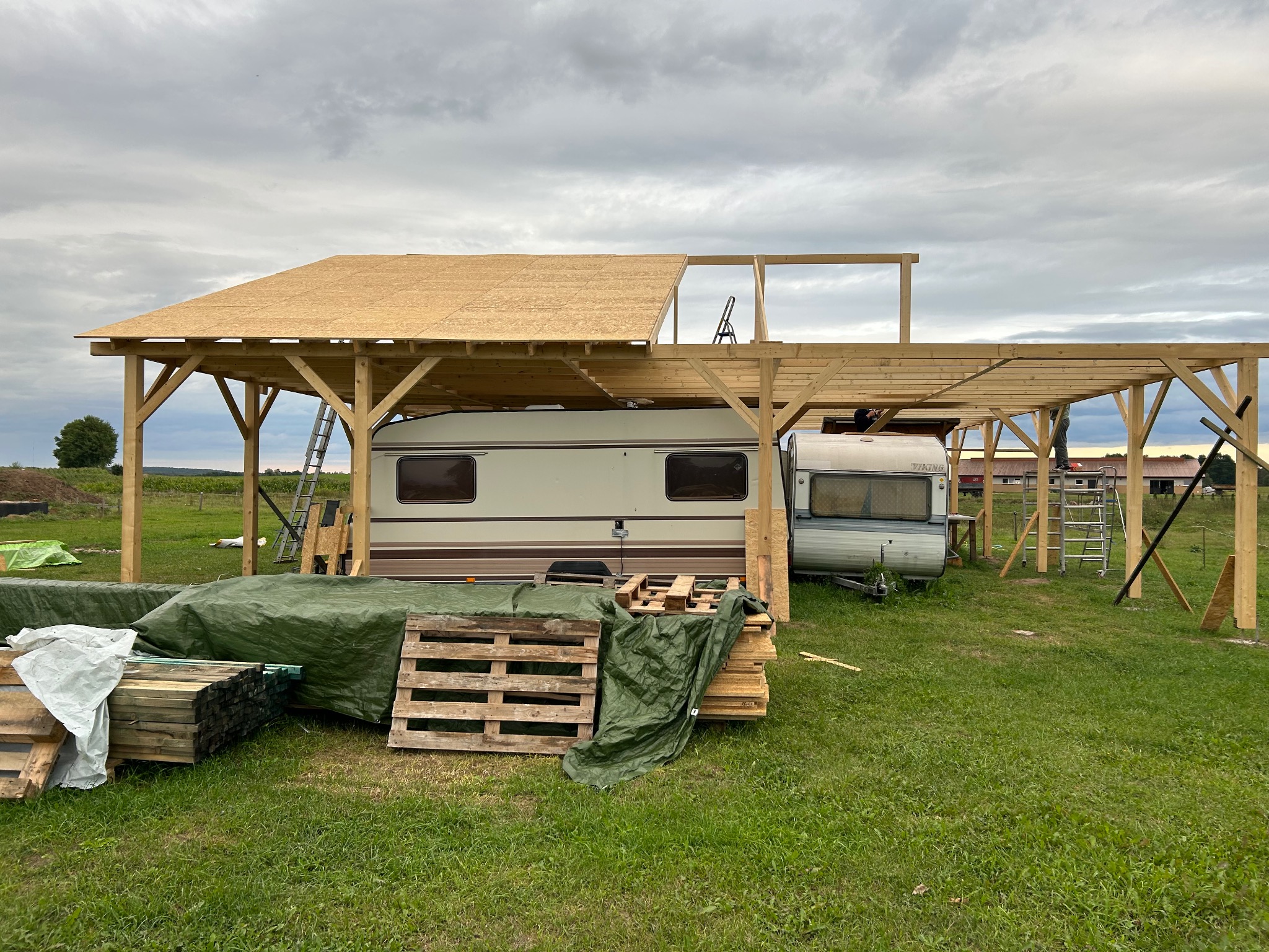 Budowa drewnianej wiaty z dachem z płyt OSB nad przyczepami kempingowymi, widoczne rusztowania, palety z drewnem i plandeka na trawie.