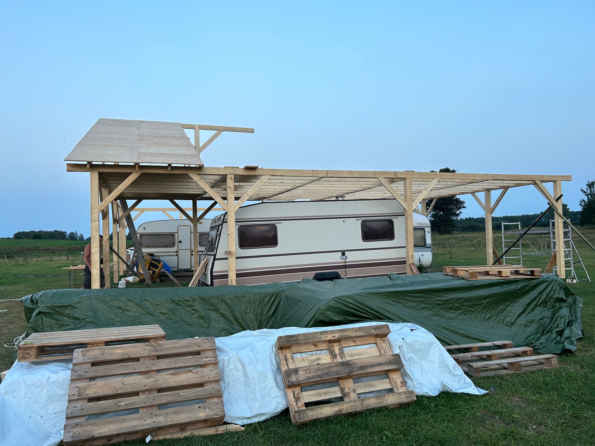 Budowa drewnianej konstrukcji z zadaszeniem obok przyczepy kempingowej, widoczne palety i plandeka ochronna w otoczeniu zielonego terenu.
