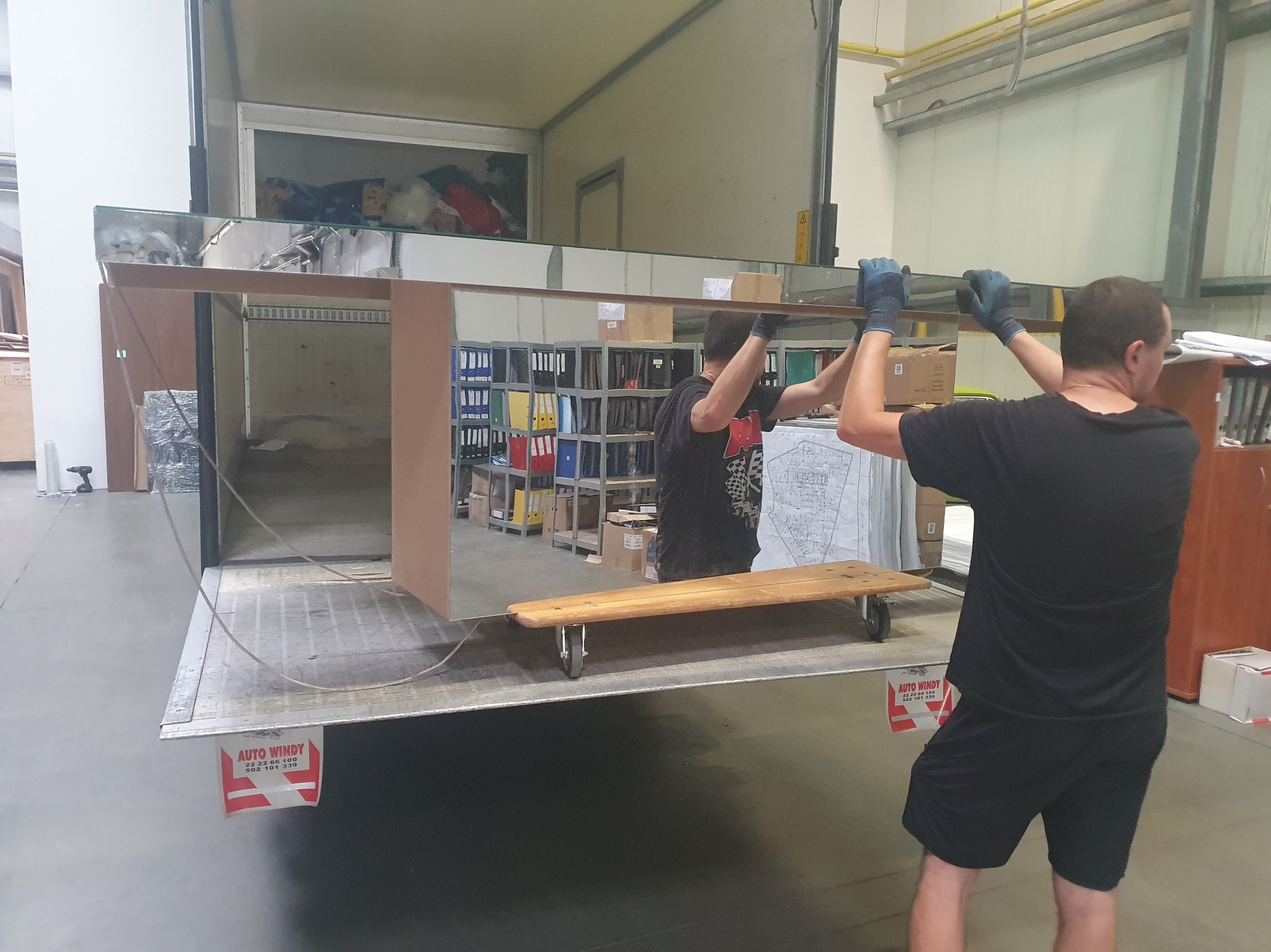 Pracownicy w rękawicach wnoszą lustro do wnętrza busa transportowego. Widoczny wózek transportowy i logo firmy na burcie. Przeprowadzka biura.