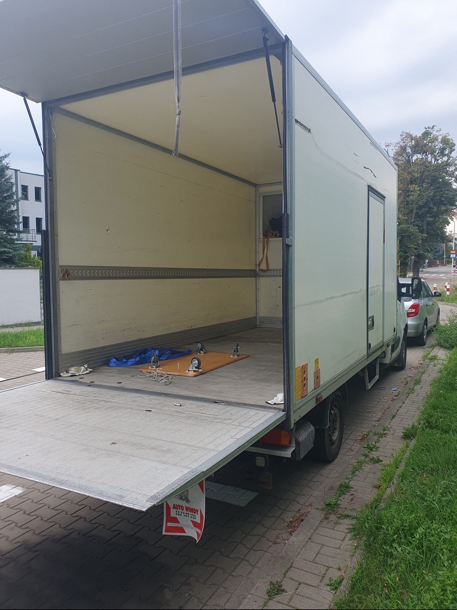 Otwarty, pusty bus przeprowadzkowy z rampą załadowczą na chodniku, obok zaparkowany samochód osobowy. Widoczne zabezpieczenia ładunku wewnątrz.