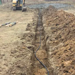 Wykop pod instalację z ułożonym kablem, widziany z góry. W tle żółta koparka i przygotowany teren. Świeżo wykopana ziemia.