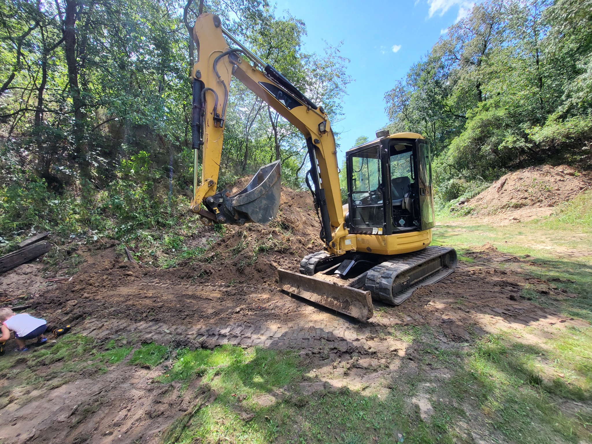 Żółta koparka na nierównym terenie z ziemią i trawą, otoczona drzewami pod błękitnym niebem. Dziecko bawi się w pobliżu. Ślady opon na ziemi.