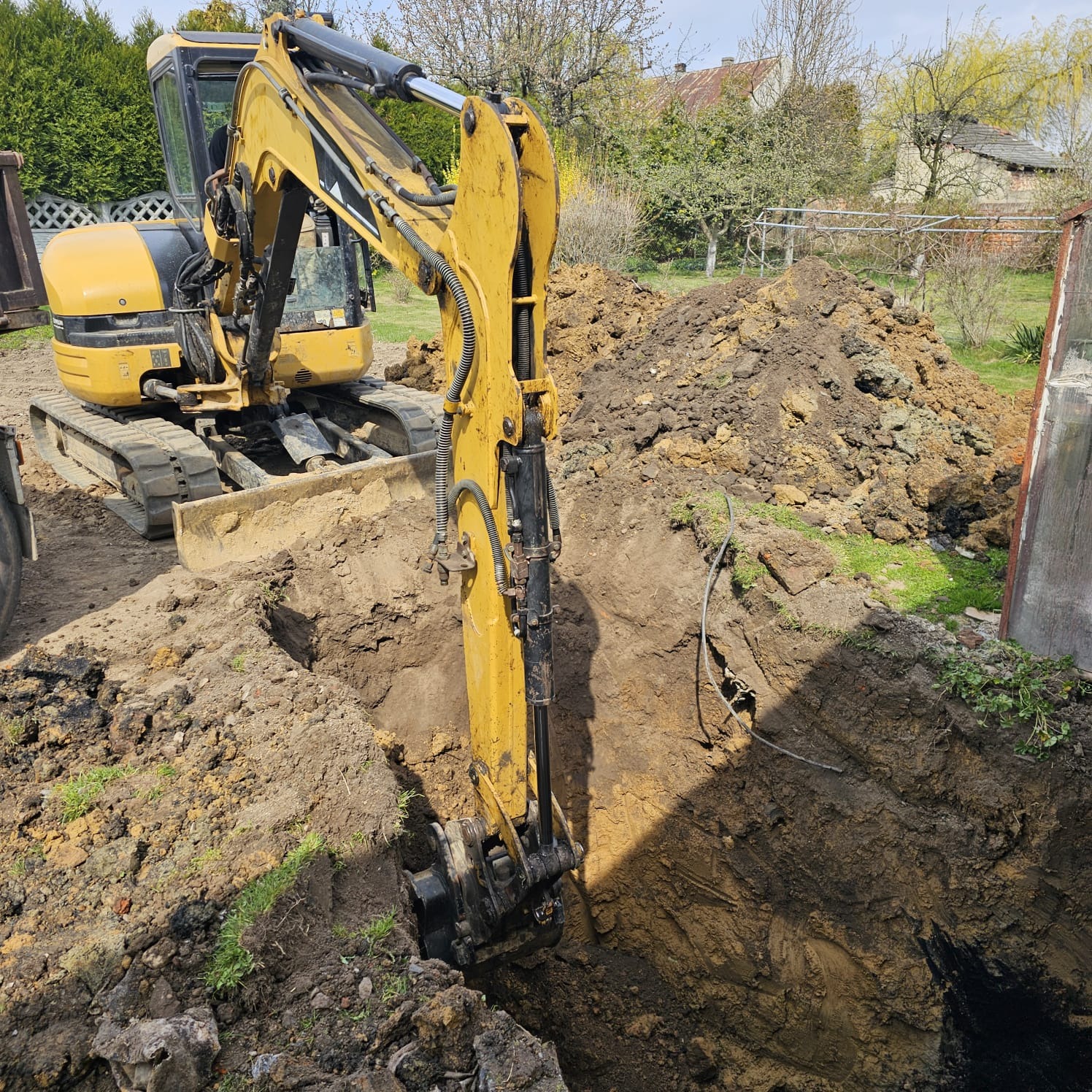 Żółta koparka w trakcie pracy na działce, wykop głębokiego rowu w ziemi. W tle widoczne budynki i drzewa.