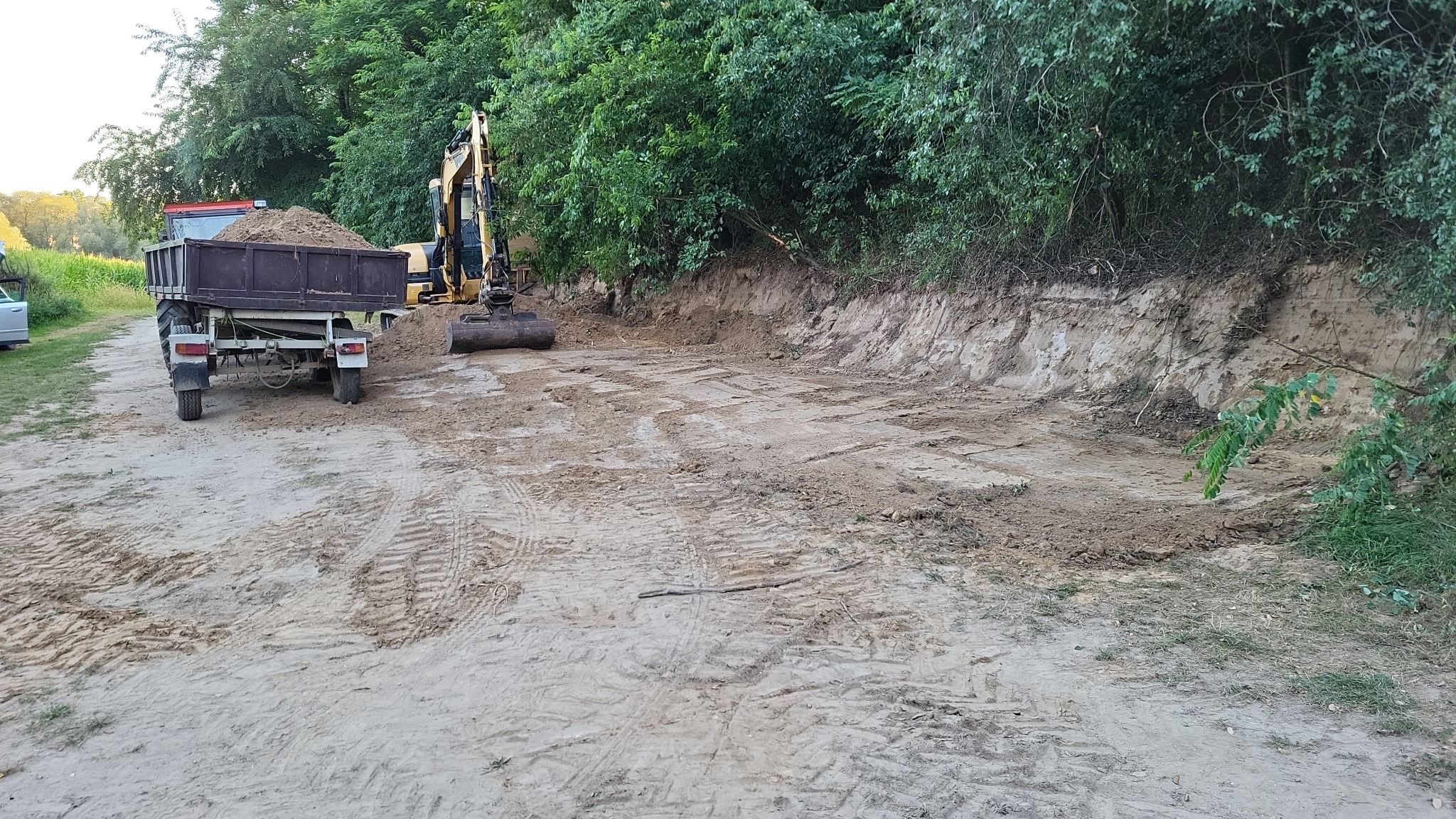 Żółta koparka i przyczepa z ziemią na wyrobisku. Ślady opon w piasku. Teren wyrównany, przygotowany pod dalsze prace. Zarośla w tle.