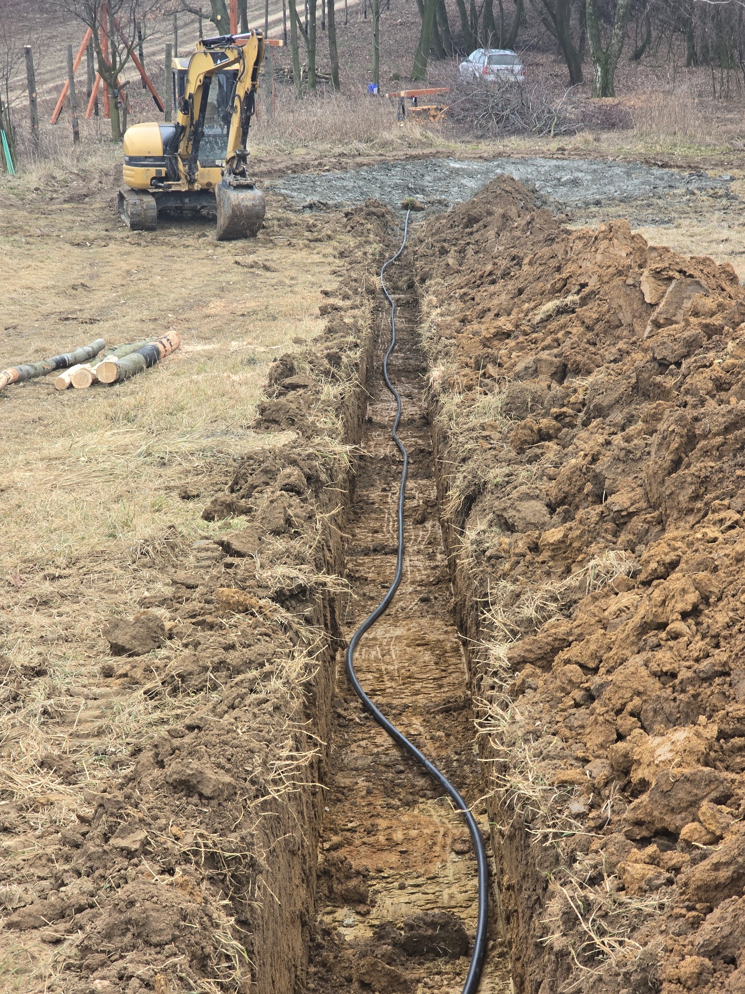 Wykop pod instalację z ułożonym kablem, widziany z góry. W tle żółta koparka i przygotowany teren. Świeżo wykopana ziemia.